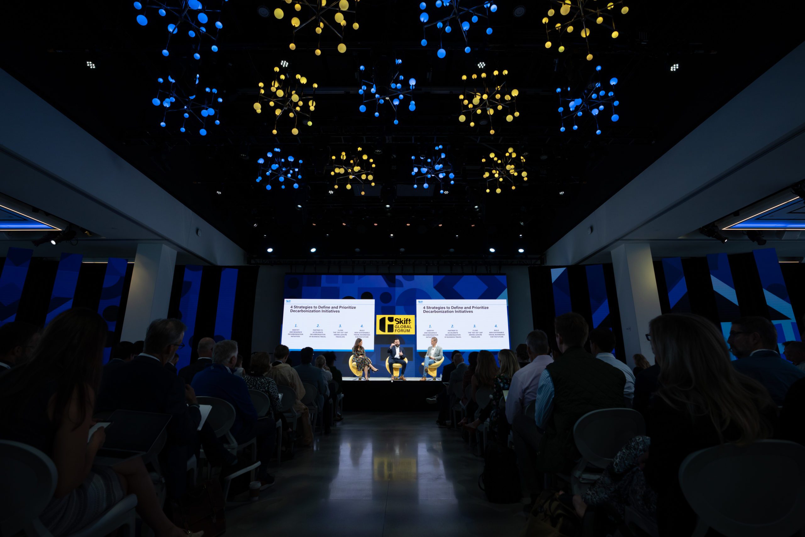 The stage of Skift Global Forum 2022 hosted in The Glasshouse in New York City venue. The image shows 3 speakers on stage in front of a large audience.