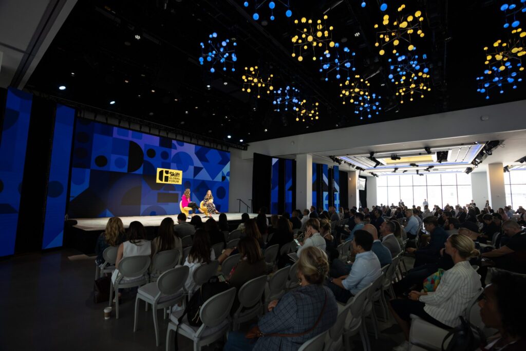 Skift Global Forum 2022 image showing 2 people on stage discussing travel industry insights in front of a large audience of attendees inside The Glasshouse venue in New York City.